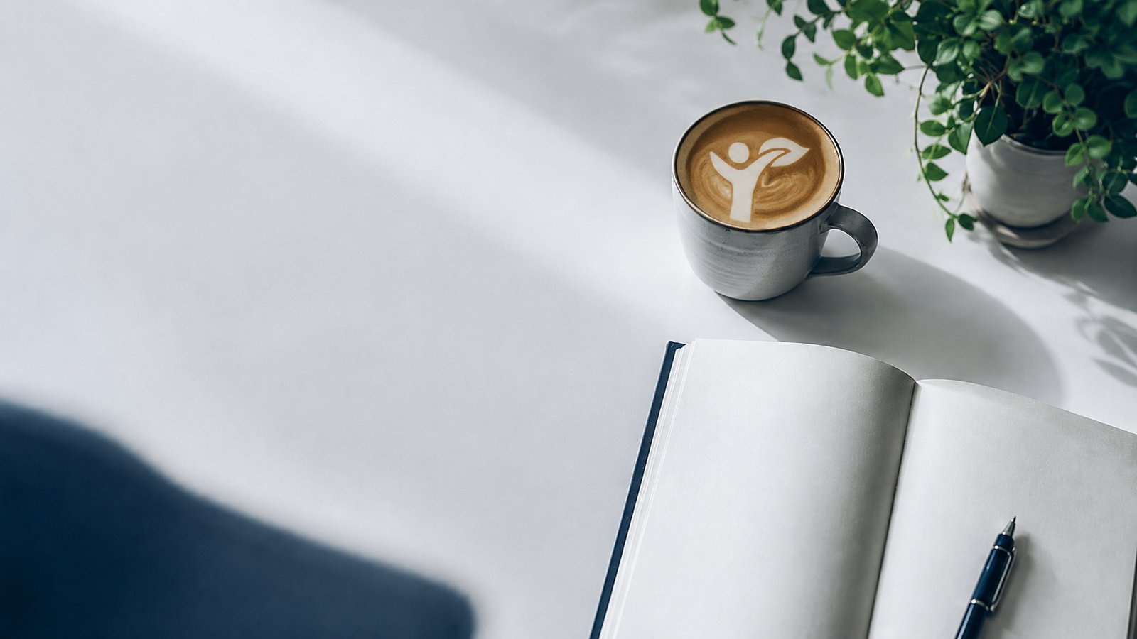 Kaffee mit VIVAME-Logo in der Latte Art, Notizbuch und Pflanze auf hellem Tisch